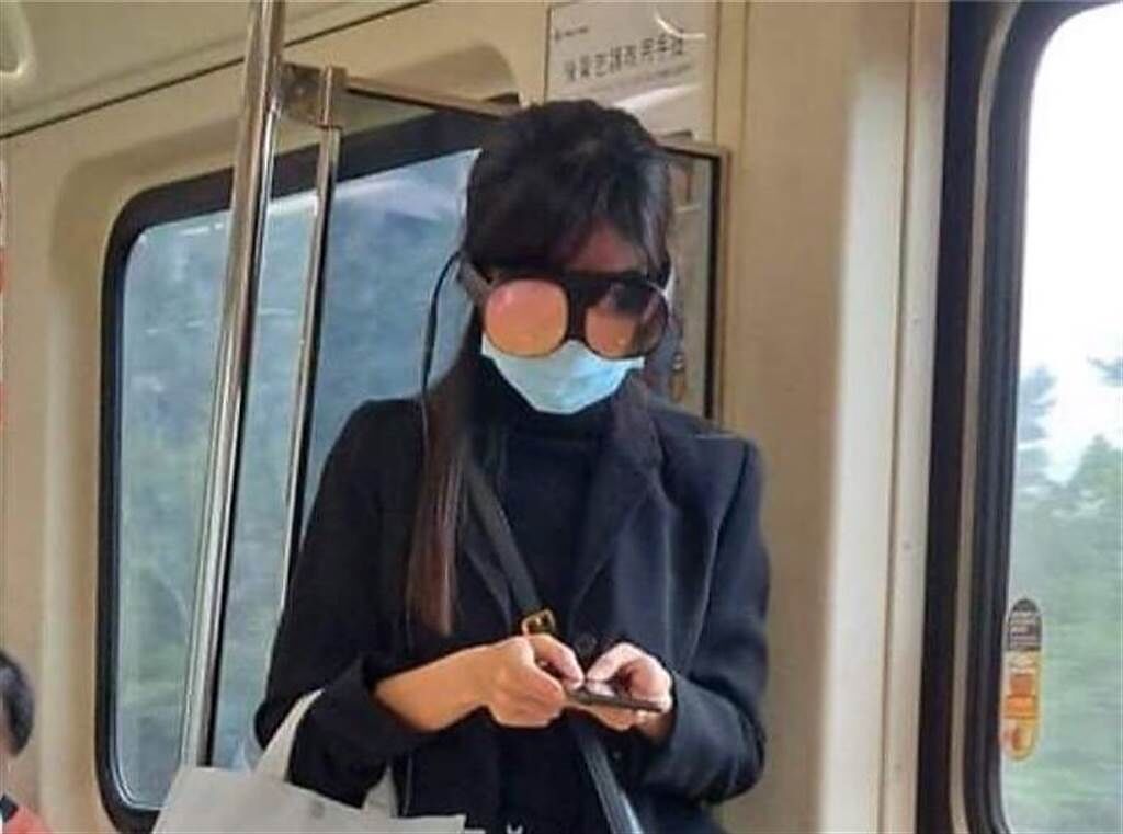 一位穿著黑色套裝配黑絲襪的女生站在捷運的門旁邊，頭上戴著碩大無比的眼鏡低著頭，專注玩手機。（圖／翻攝自爆廢公社）